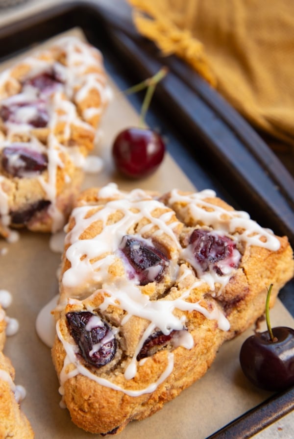 Baking sheet with large cherry scones and fresh cherries to the side with a golden napkin in the background.
