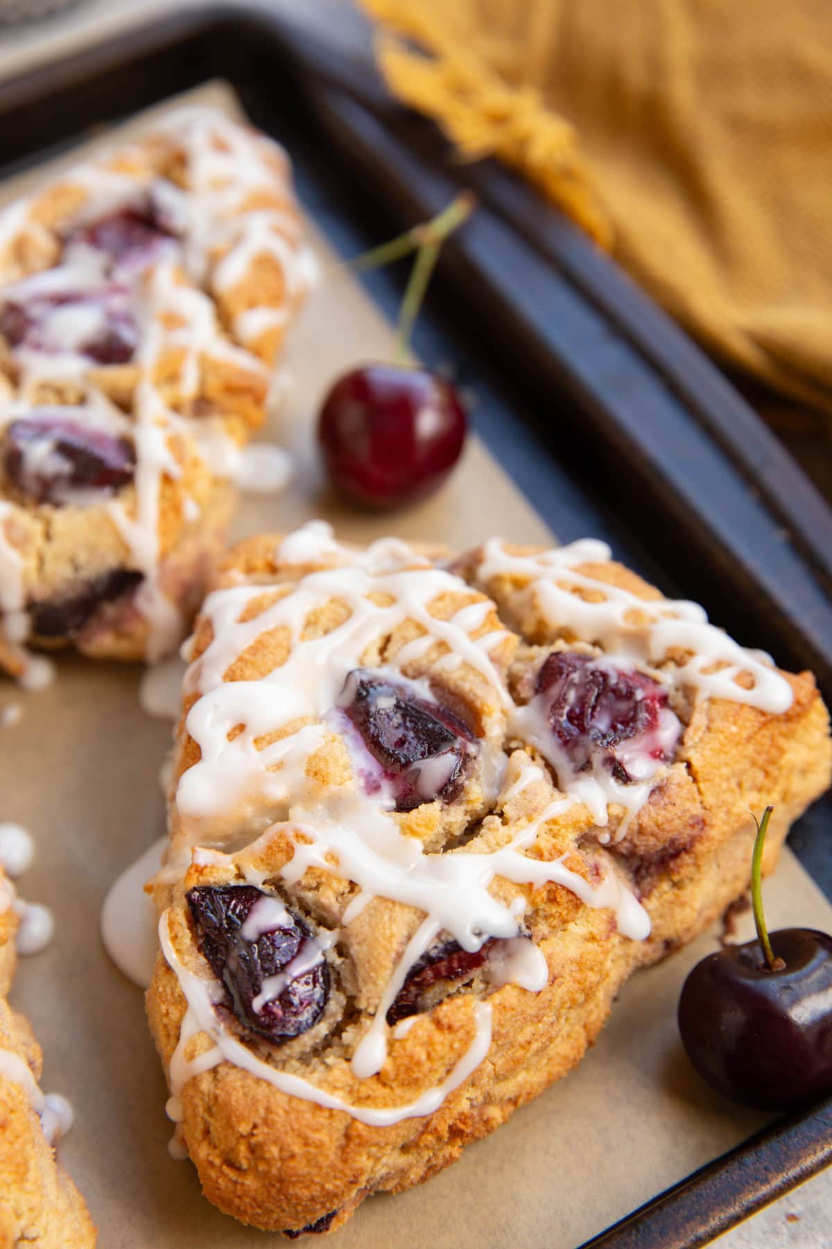 Baking sheet with large cherry scones and fresh cherries to the side with a golden napkin in the background.