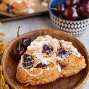 Cherry scone with glaze on top on a wooden plate with two fresh cherries, a bowl of fresh cherries in the background, and the baking sheet with the rest of the cherries.