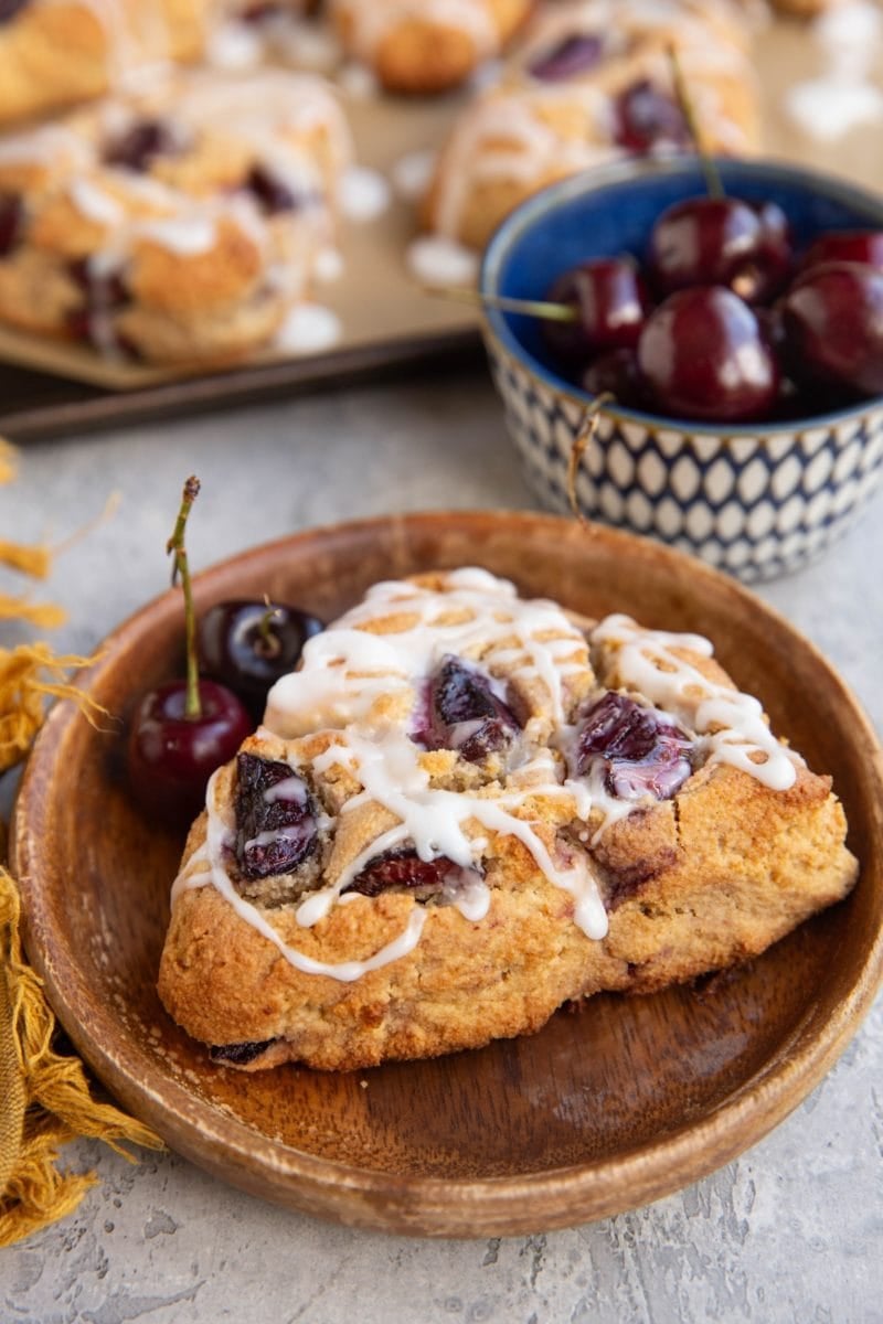 Cherry scone with glaze on top on a wooden plate with two fresh cherries, a bowl of fresh cherries in the background, and the baking sheet with the rest of the cherries.