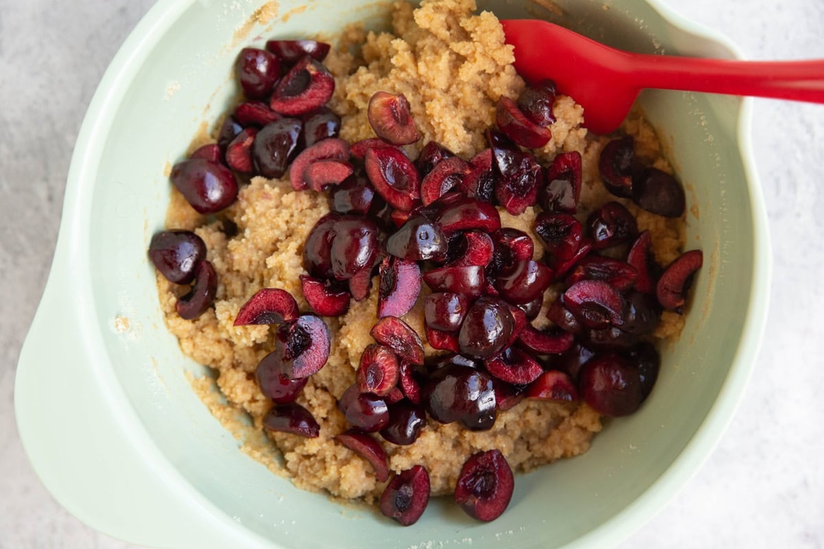 Scone dough in a mixing bowl with chopped cherries on top and a rubber spatula to mix them in.