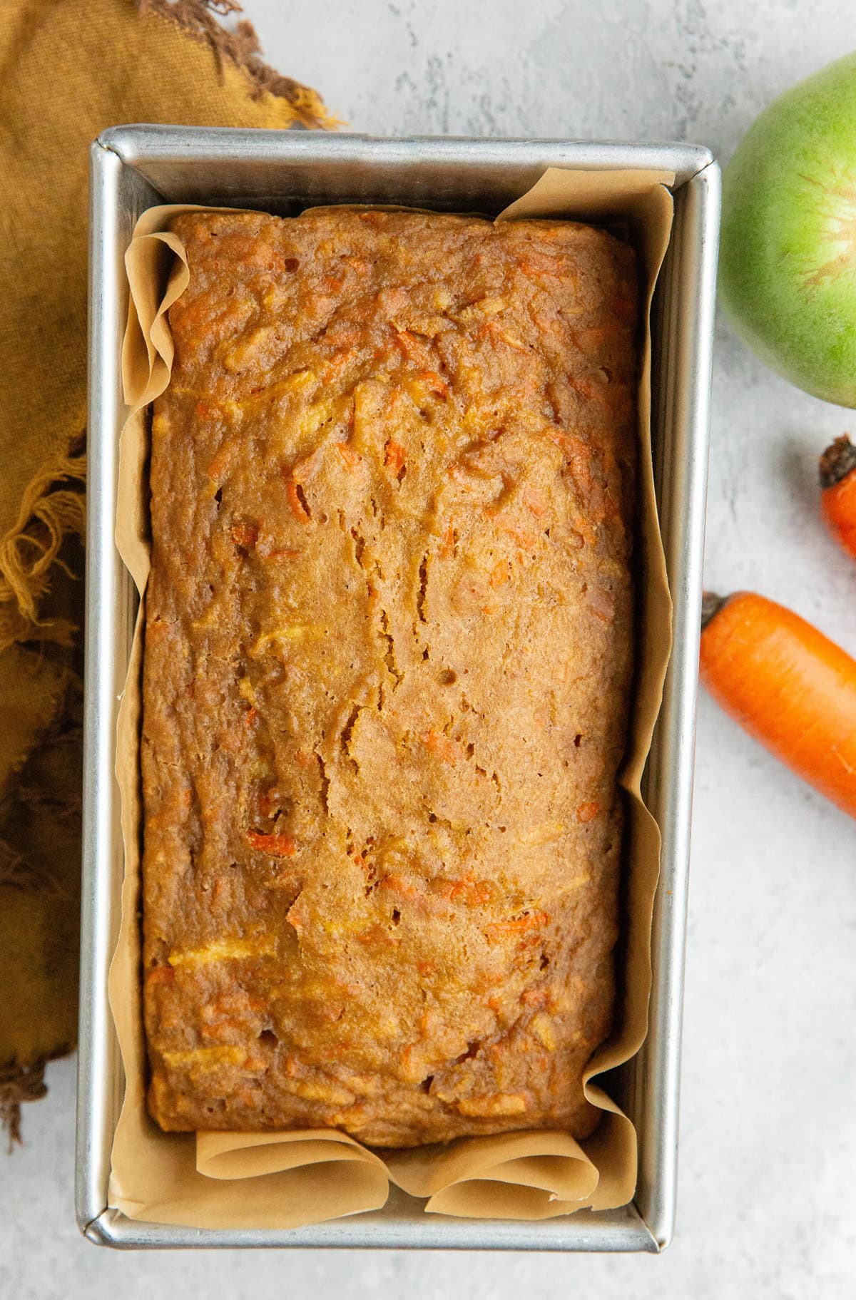 Loaf pan with carrot apple oat bread inside with fresh carrots and an apple to the side.