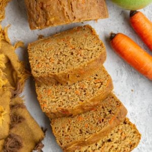Loaf of carrot apple bread cut into slices with fresh carrots and a green apple to the side.