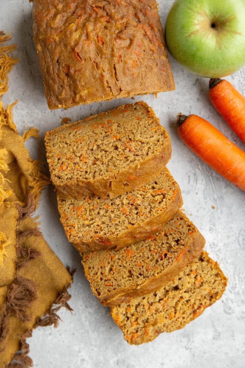 Loaf of carrot apple bread cut into slices with fresh carrots and a green apple to the side.
