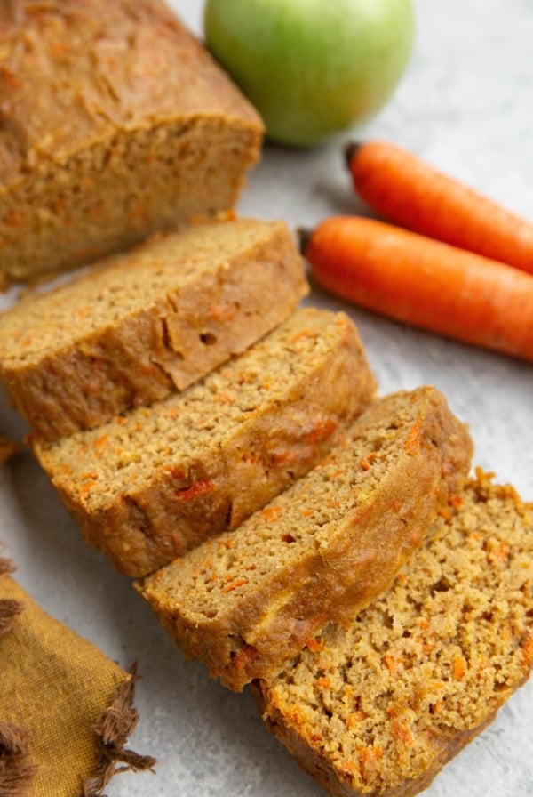 Loaf of carrot apple bread cut into slices with fresh carrots and a green apple in the background.