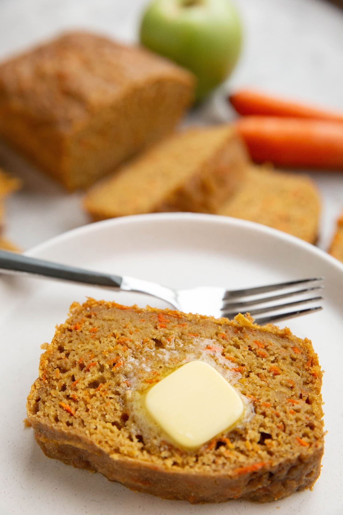 Slice of carrot apple bread on a plate with a pat of melted butter on top and a fork to the side with the rest of the loaf in the background, cut into slices.
