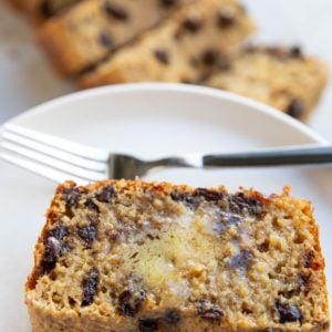 Thick slice of chocolate chip bread on a white plate with melted butter on top, a fork for eating, and the rest of the loaf sliced in the background.