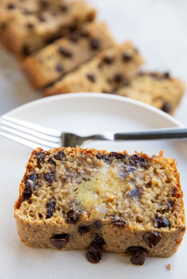Thick slice of chocolate chip bread on a white plate with melted butter on top, a fork for eating, and the rest of the loaf sliced in the background.