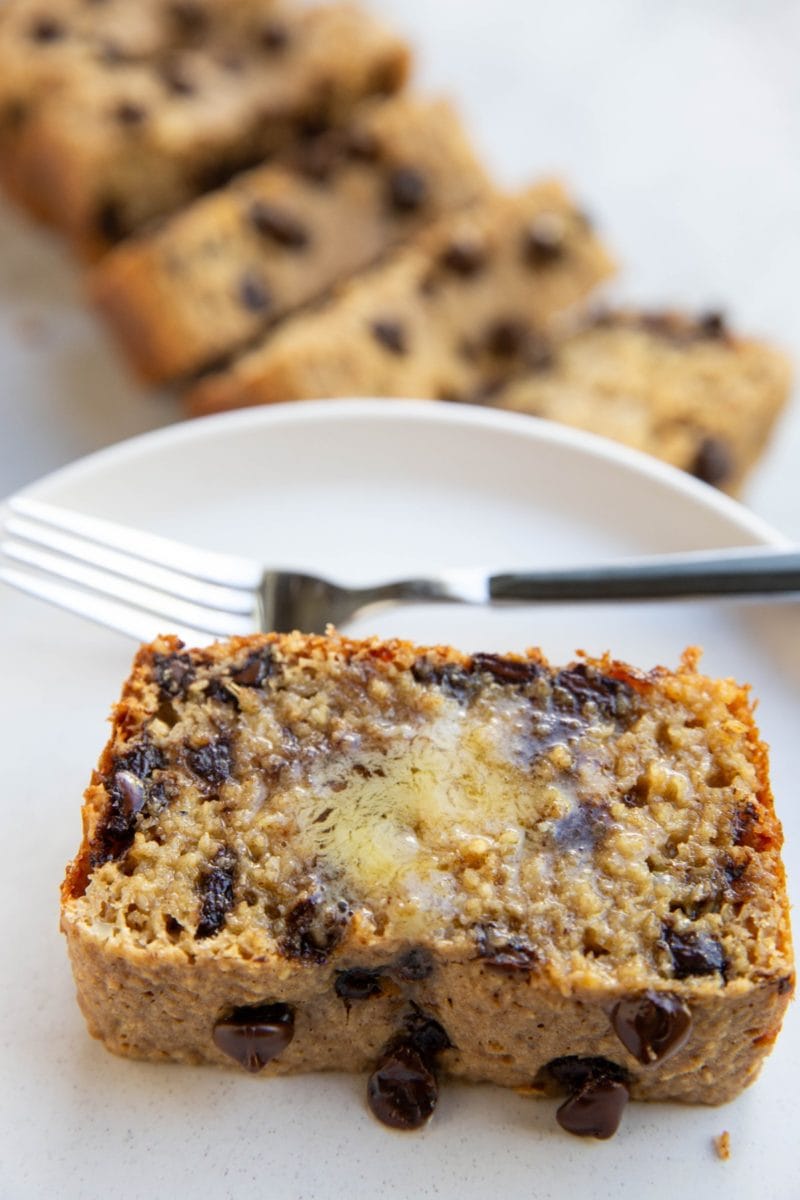 Thick slice of chocolate chip bread on a white plate with melted butter on top, a fork for eating, and the rest of the loaf sliced in the background.