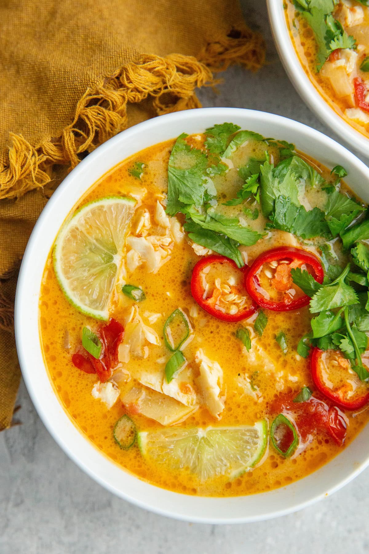 Bowl full of Coconut Lime Fish Soup sprinkled with green onions, with lime wedges, sliced chili peppers, and fresh cilantro on top.