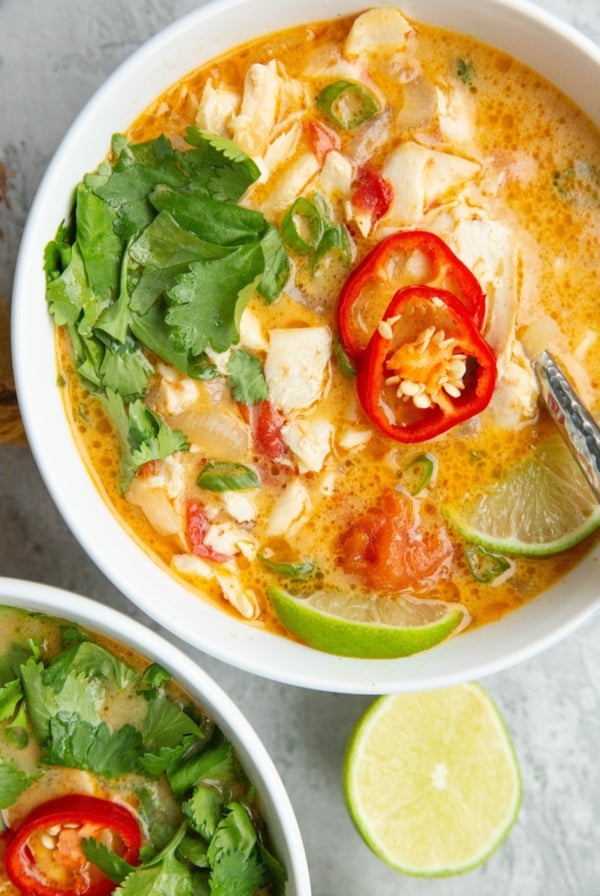 Coconut Lime fish soup in two large white bowls with chili peppers, cilantro, and lime wedges on top, ready to serve.