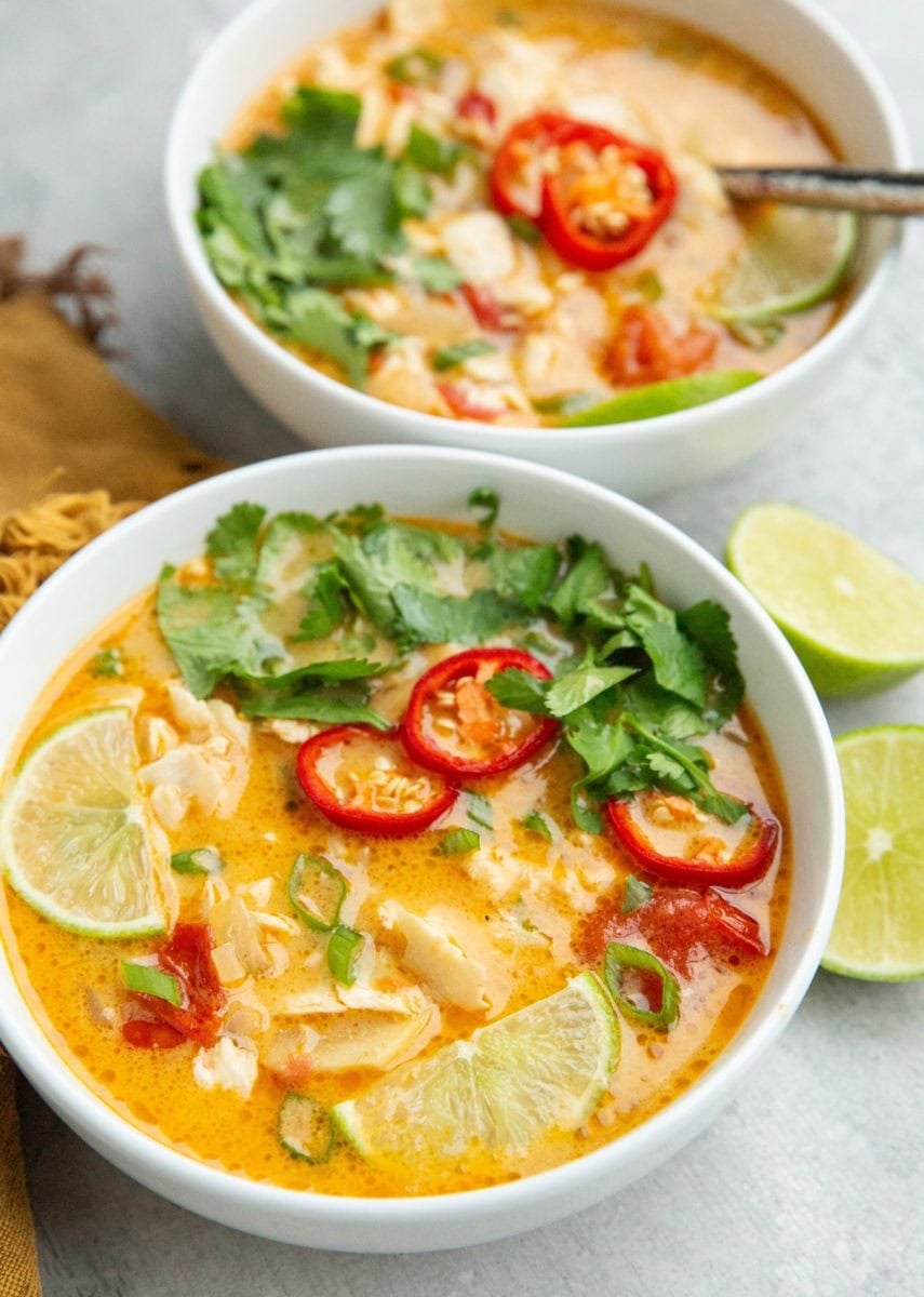 Two bowls of coconut lime fish soup with lime wedges, cilantro, and red chili peppers on top with a cut lime to the side and a golden napkin.