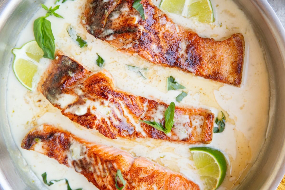 Three golden brown seared salmon filets in a stainless steel skillet full of creamy coconut sauce with fresh limes and basil. Ready to serve.