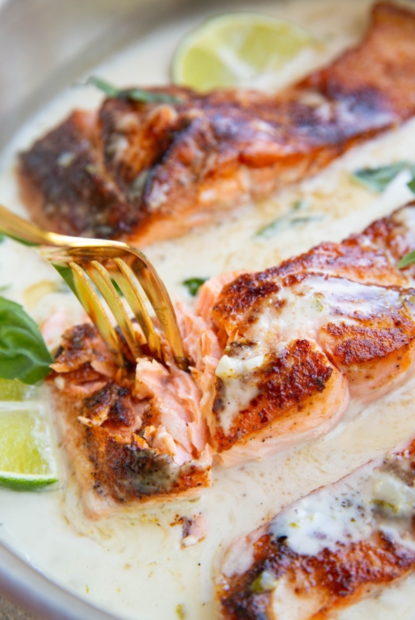 Three salmon filets cooked in a skillet with a fork digging into one of them so you can see the juicy flaky texture. The salmon is surrounded by creamy coconut sauce.