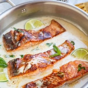 Stainless steel skillet full of three salmon filets in a creamy coconut milk sauce with lime and basil.