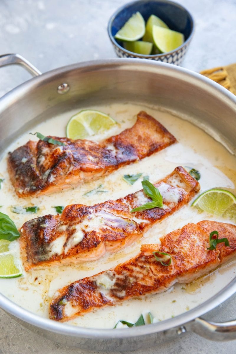 Stainless steel skillet full of three salmon filets in a creamy coconut milk sauce with lime and basil.