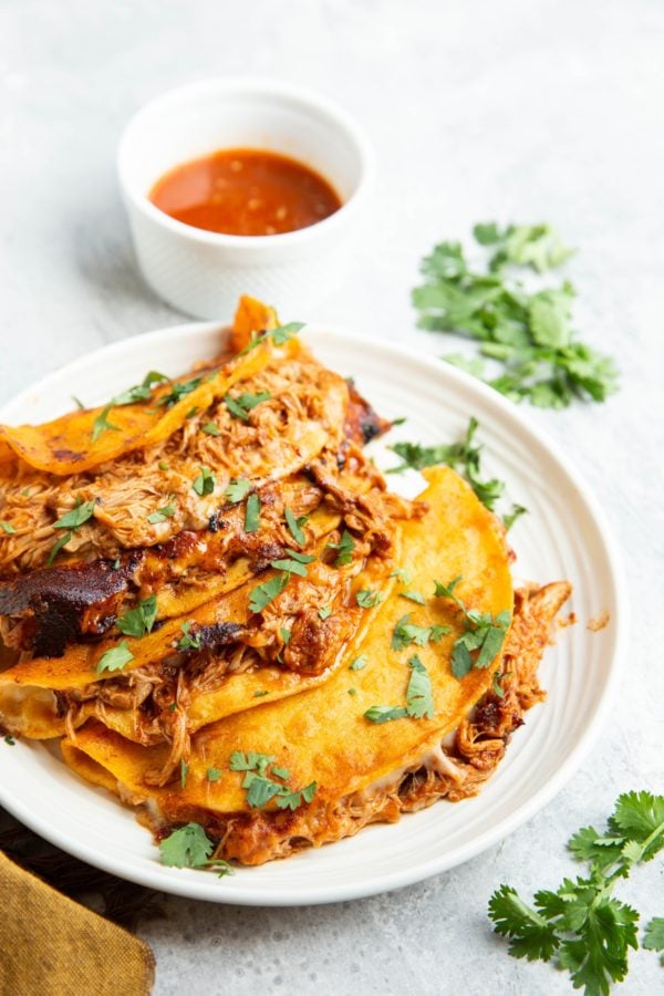 Four chicken birria tacos on a plate with a bowl of consommé in the background and fresh cilantro to the side.