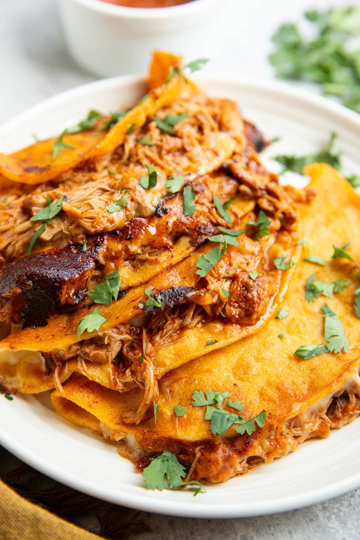Four crispy corn tortilla tacos on a plate with melted cheese and shredded chicken. Everything sprinkled with fresh cilantro and a bowl of sauce in the background.