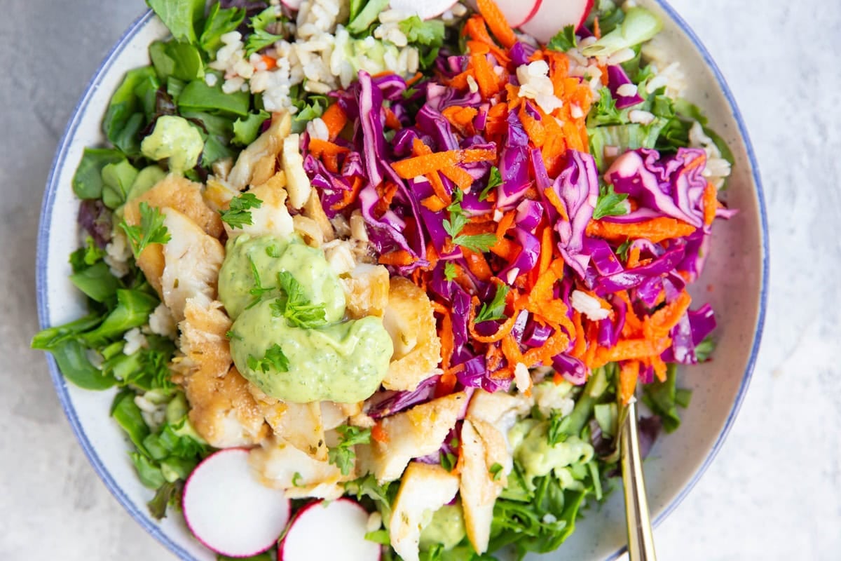 Bowl full of greens, rice, fish, cabbage slaw, and avocado sauce for a fish taco bowl.