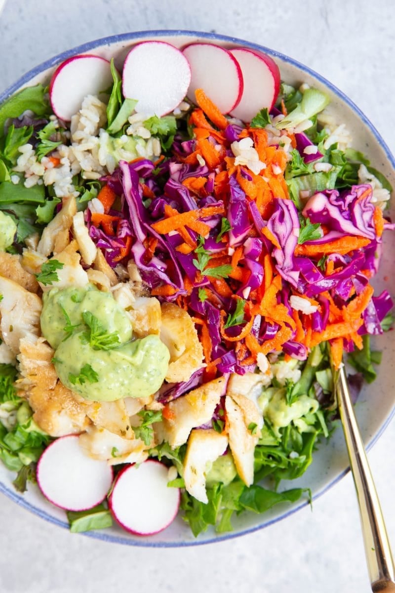 Large blue-rimmed bowl full of greens, rice, cabbage and carrot slaw, fresh cooked flaky fish, and avocado sauce with a spoon to the side. The ultimate fish taco bowl.