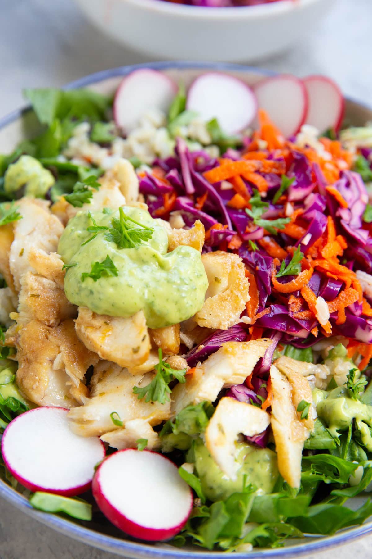 Cooked fish with creamy avocado sauce on top of it on a bed of greens with cabbage slaw to make the most amazing fish taco bowls.