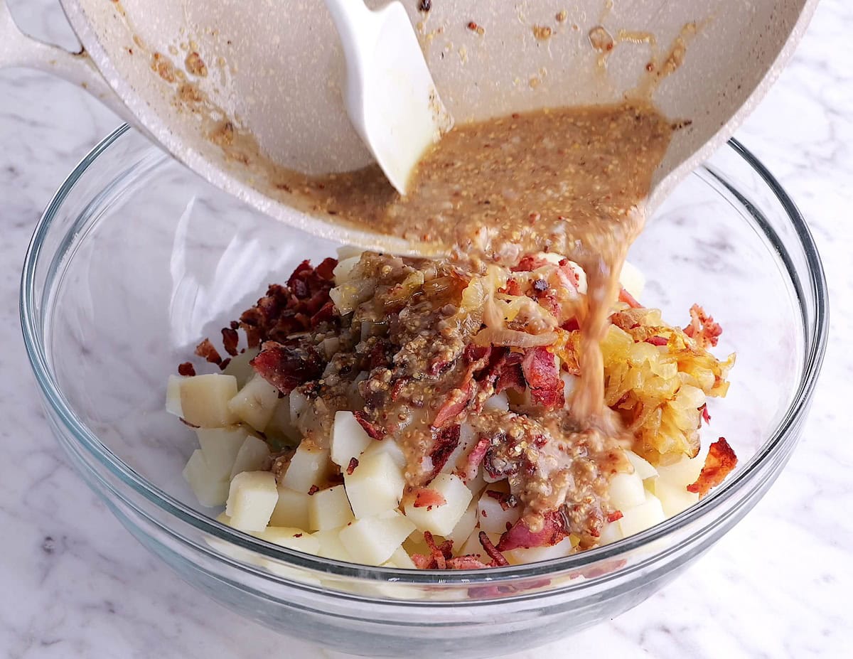 Pouring potato salad dressing into a bowl with potatoes, bacon, and caramelized onions.
