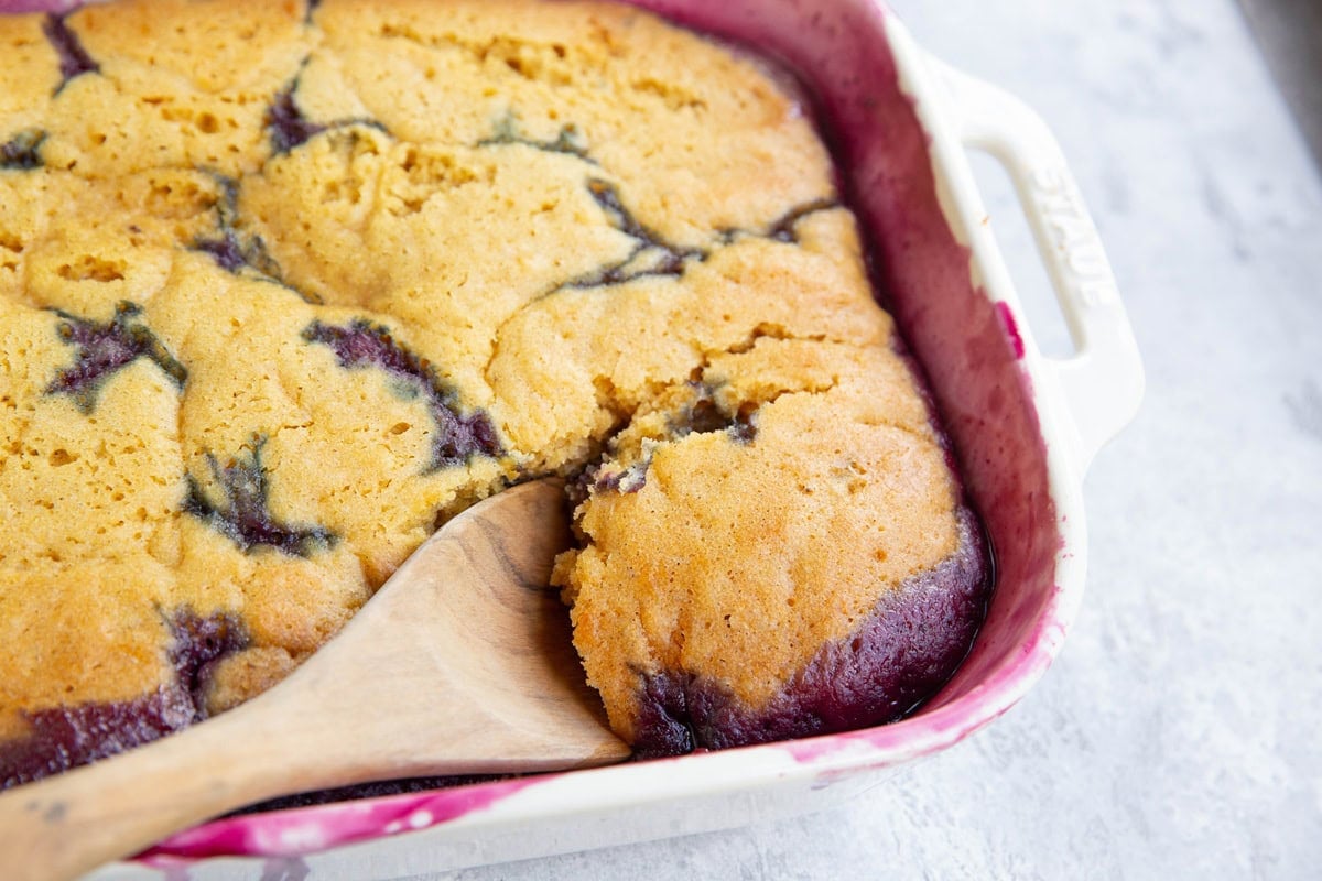 Casserole dish full of berry cobbler with a cake topping instead of a biscuit topping and a wooden spoon scooping out some of the cobbler to serve.