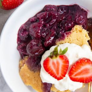 White bowl full of gluten-free berry cobbler with whipped cream on top and fresh strawberries and a golden spoon to the side, ready to serve.