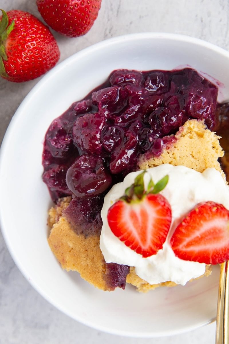 White bowl full of gluten-free berry cobbler with whipped cream on top and fresh strawberries and a golden spoon to the side, ready to serve.