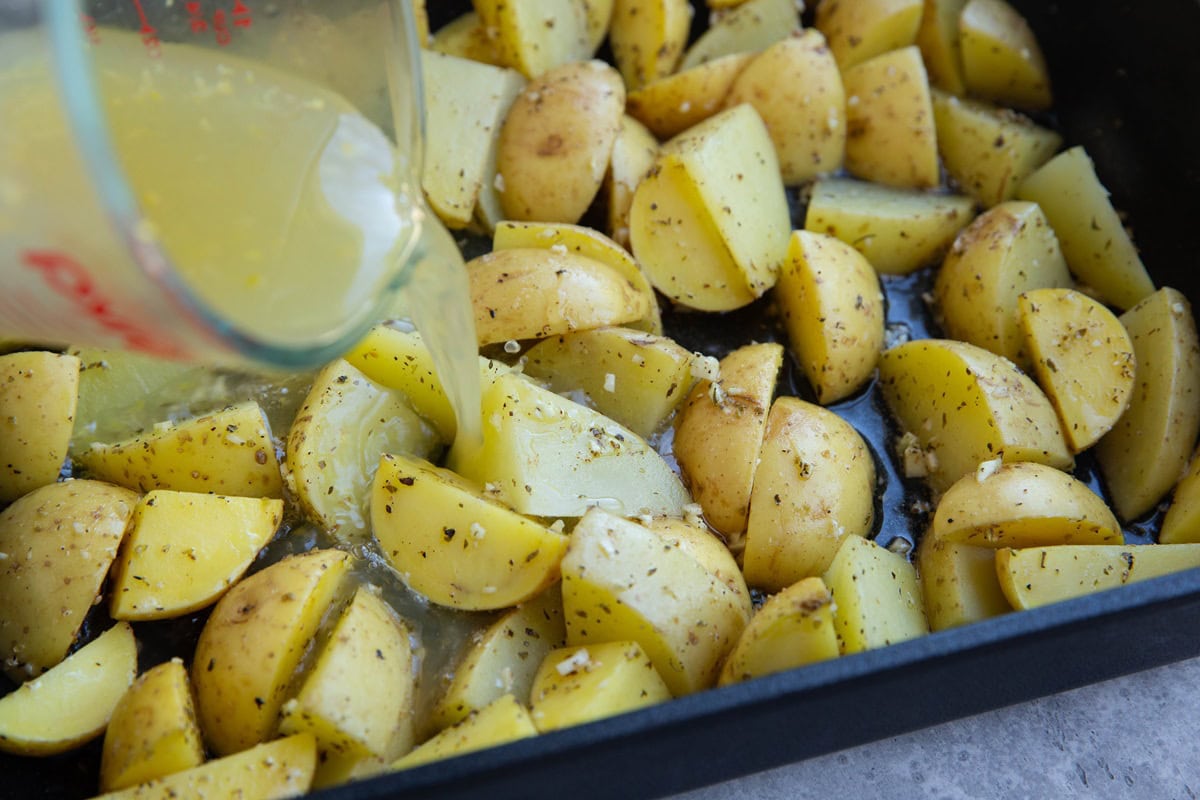 Pouring lemon chicken broth over roasted potatoes in a baking dish to continue baking.