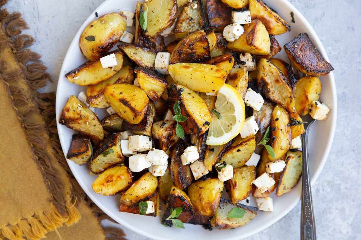 White serving plate with roasted potatoes on top with feta cheese cubes, fresh oregano, and fresh lemon slices.