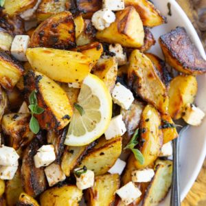 White plate full of Greek lemon roasted potatoes with sliced lemon, feta cheese, and oregano on top.