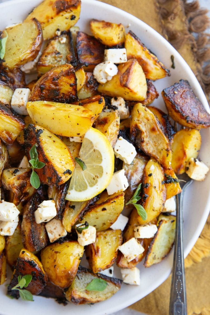 White plate full of Greek lemon roasted potatoes with sliced lemon, feta cheese, and oregano on top.