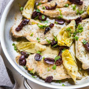 Stainless steel skillet full of chicken breasts with artichoke hearts, kalamata olives, and sun-dried tomatoes.