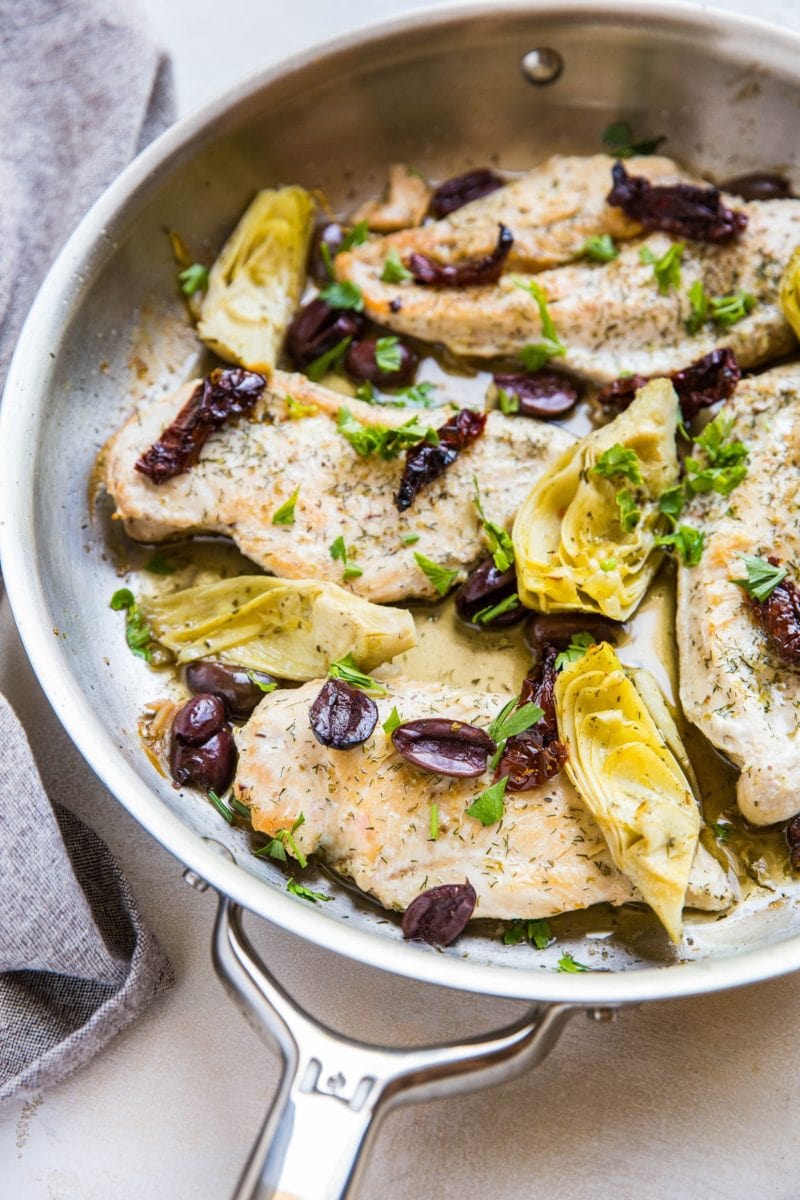 Stainless steel skillet full of chicken breasts with artichoke hearts, kalamata olives, and sun-dried tomatoes.
