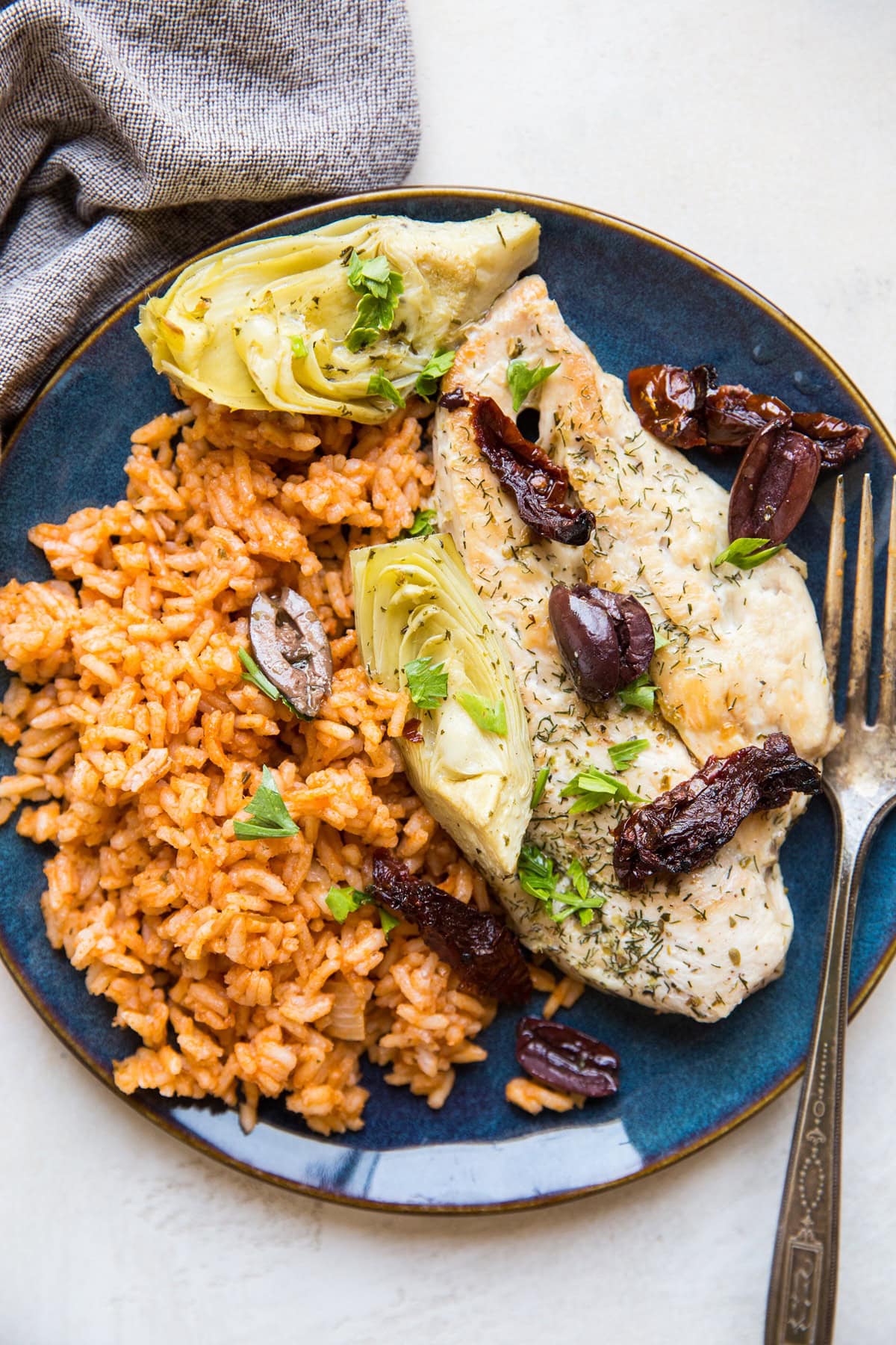Blue plate of chicken breast with artichoke hearts, olives, and sun-dried tomatoes on top, next to Spanish rice for a wholesome meal.