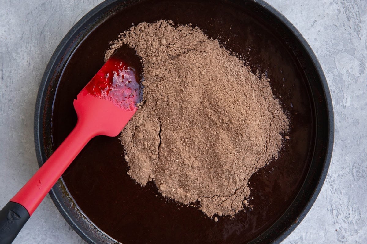 Dry ingredients for brownies on top of the wet ingredients in a skillet with a rubber spatula ready to stir.