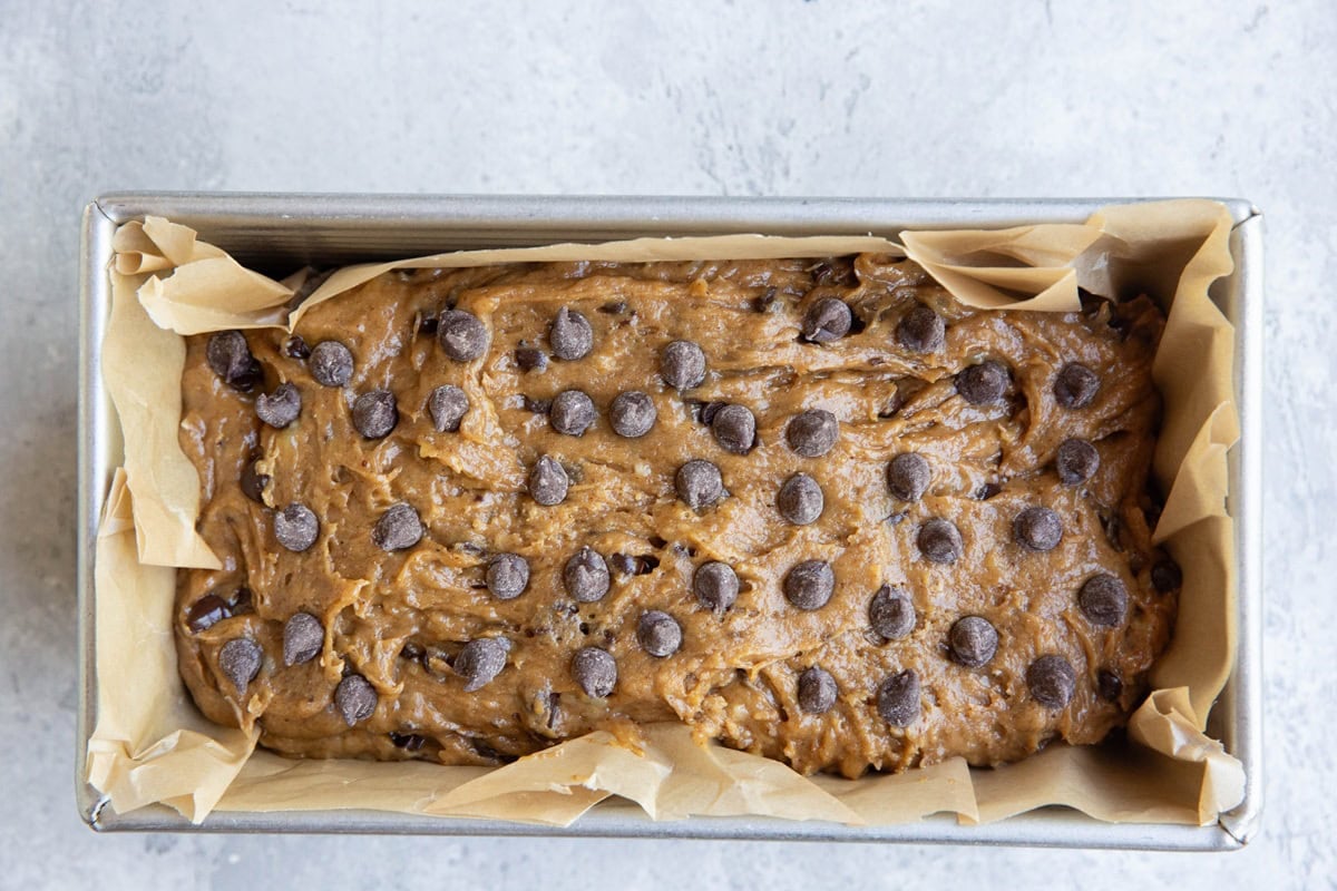 banana bread batter in a loaf pan lined with parchment paper with chocolate chips sprinkled on top, ready to go into the oven.