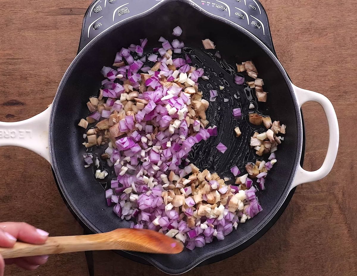 Red onion and mushrooms sautéing in a skillet with a wooden spoon stirring the contents.