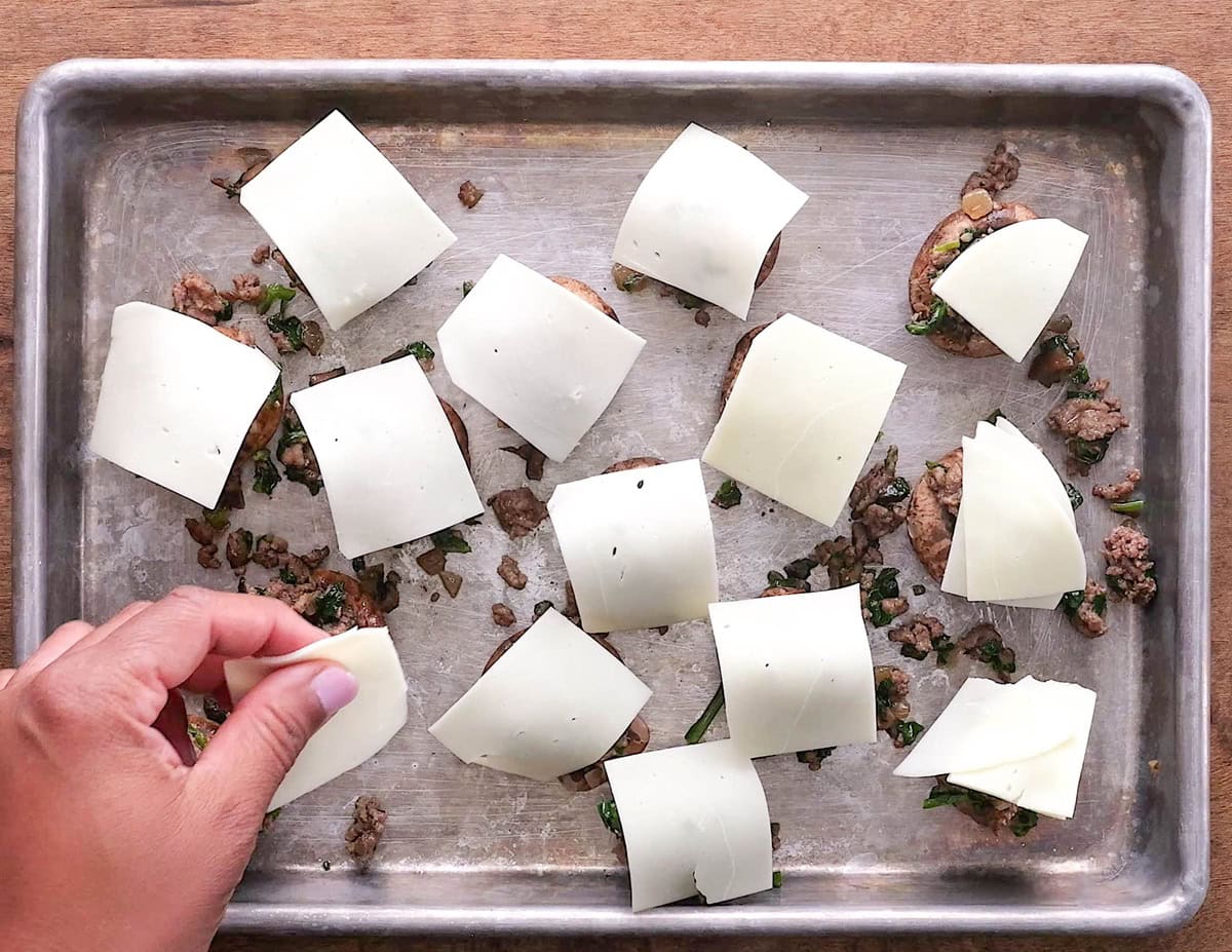 Hand placing sliced cheese on top of the meat stuffed mushrooms.