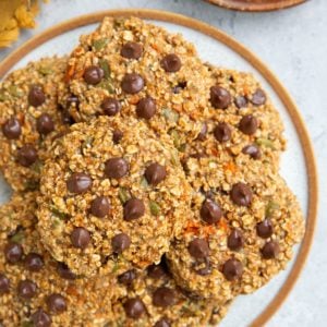 Plate of sweet potato oatmeal cookies with chocolate chips and a wooden plate with one cookie to the side and a golden napkin.