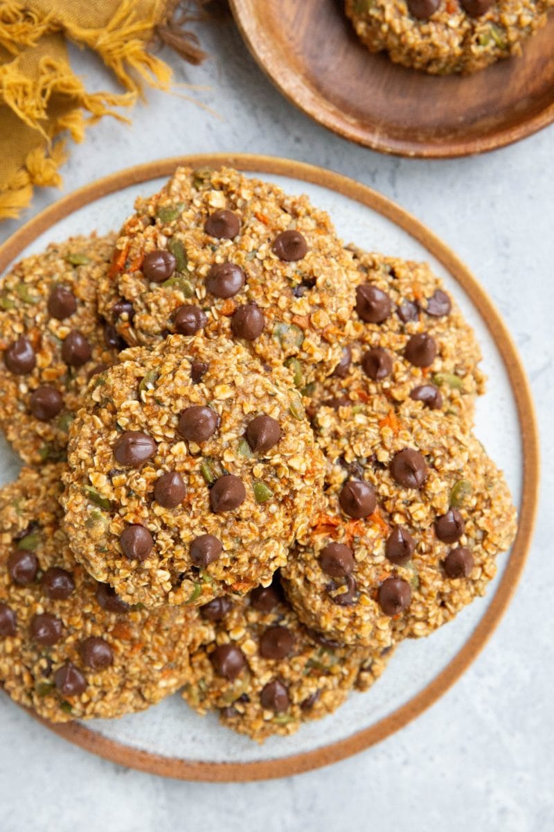 Plate of sweet potato oatmeal cookies with chocolate chips and a wooden plate with one cookie to the side and a golden napkin.