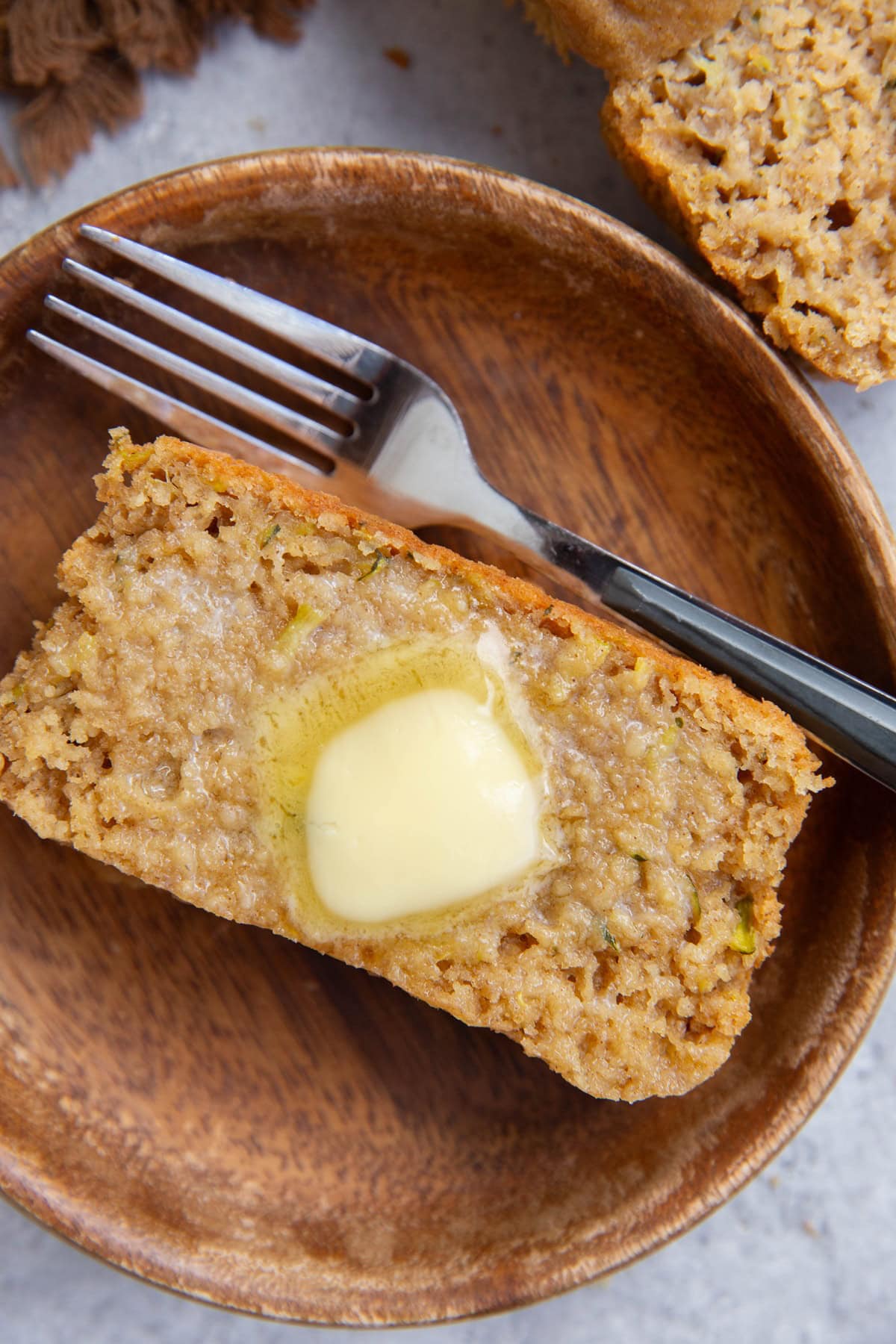 Slice of zucchini bread on a wooden plate with butter melted into the slice and a fork to the side, ready to eat.