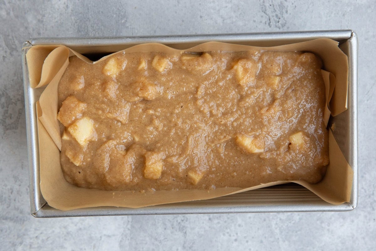 Loaf pan lined with parchment paper with apple bread batter inside, ready to be baked.