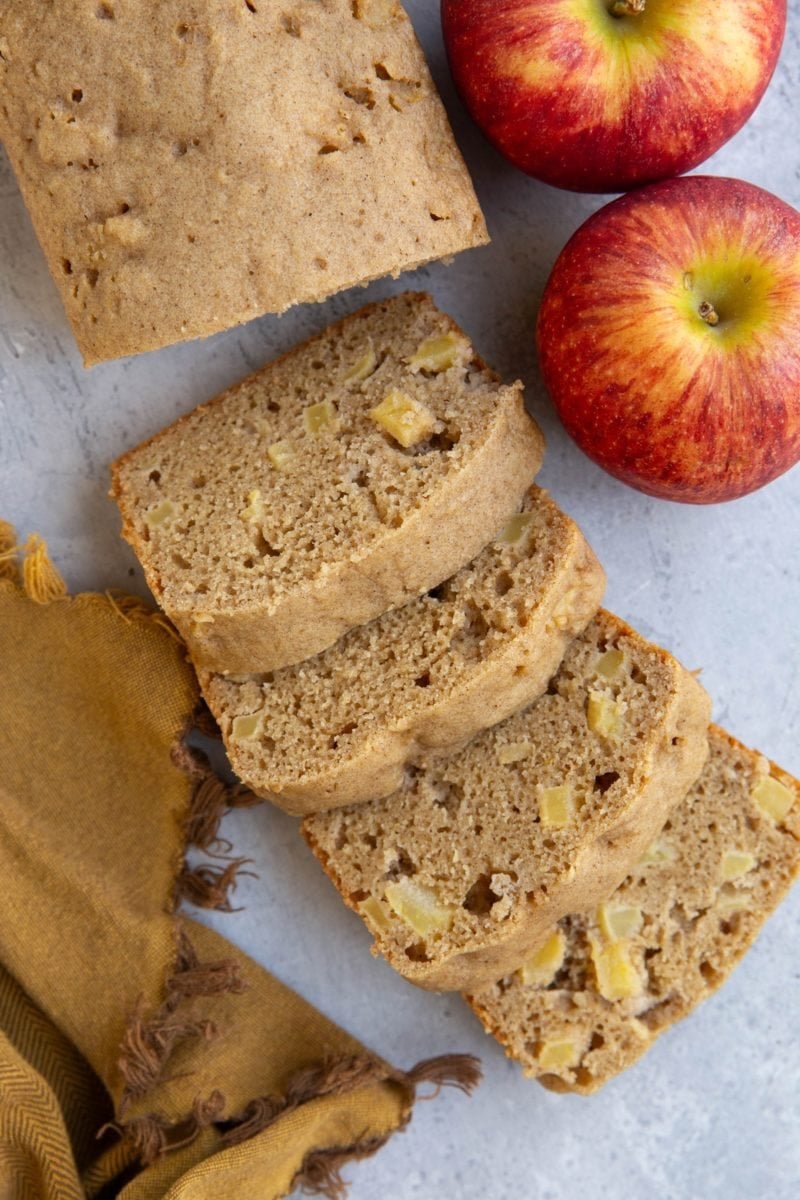 Loaf of apple bread cut into slices with two fresh apples to the side.
