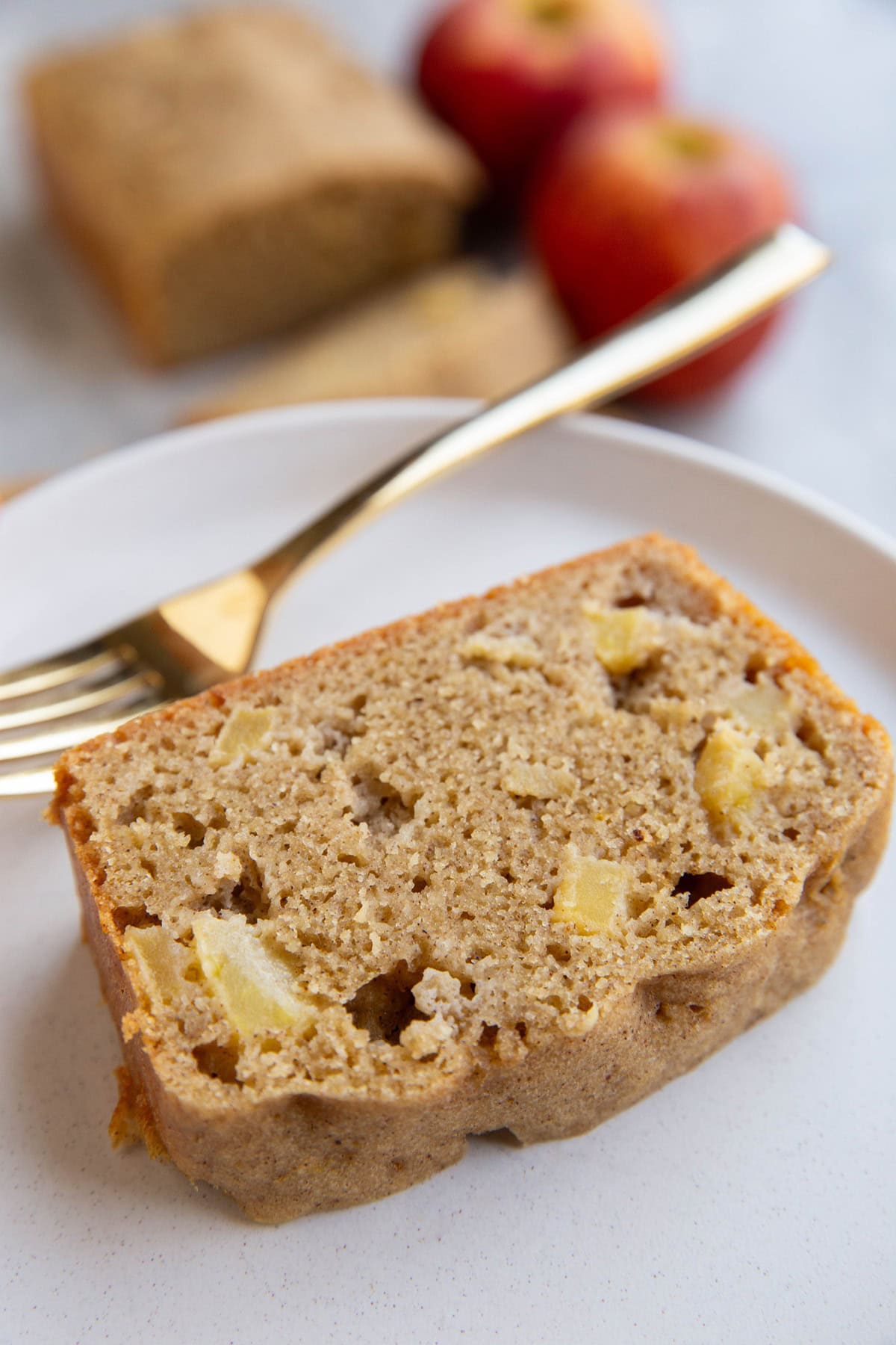 Slice of apple bread on a white plate with a golden fork with the rest of the loaf of apple bread in the background and two fresh apples.