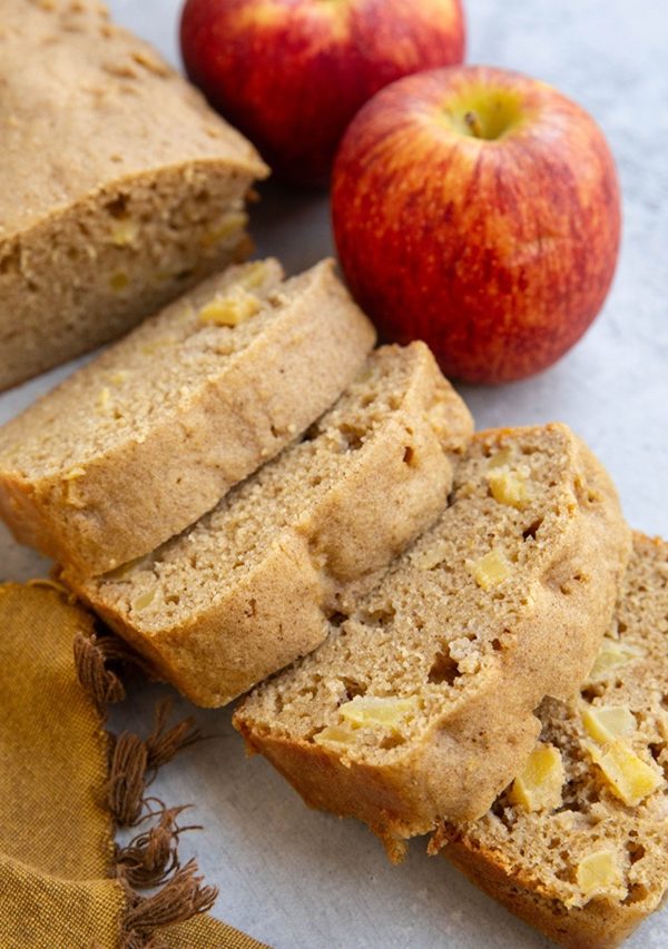 Loaf of apple bread cut into slices with fresh chunks of apples throughout the bread and fresh apples in the background.