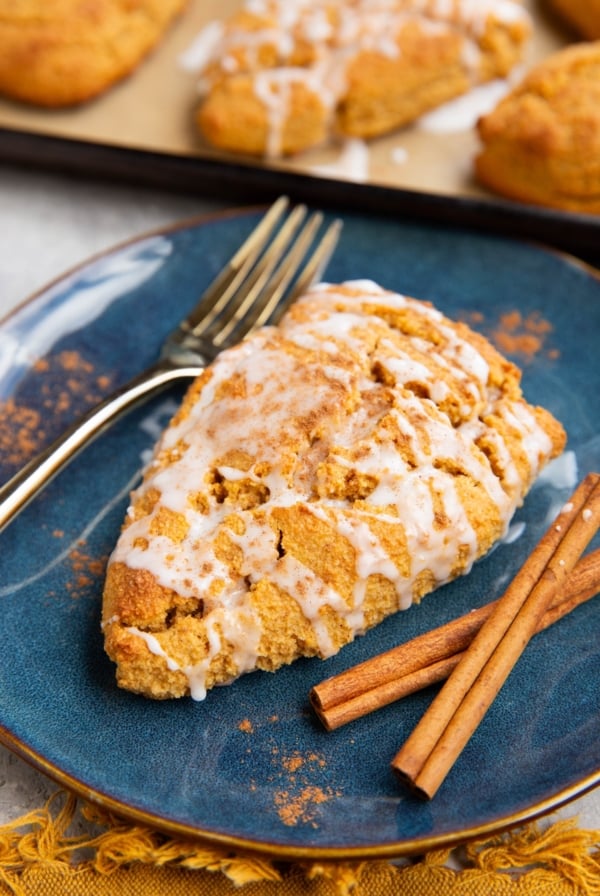 Pumpkin scone on a blue plate covered with glaze and sprinkled with pumpkin pie spice with cinnamon sticks to the side and a baking sheet of scones in the background.