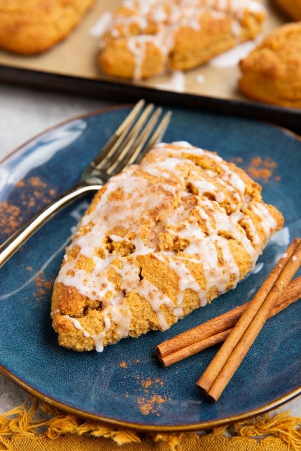Pumpkin scone on a blue plate covered with glaze and sprinkled with pumpkin pie spice with cinnamon sticks to the side and a baking sheet of scones in the background.