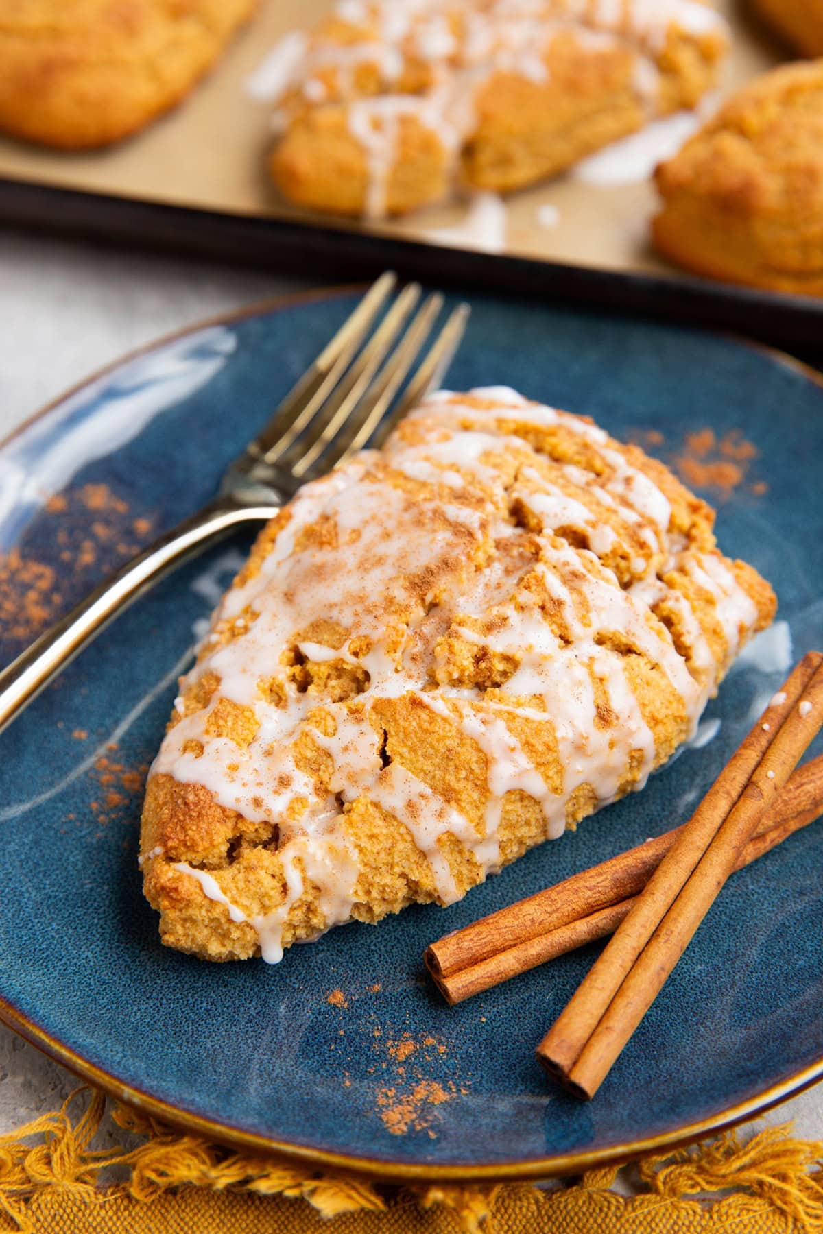 Pumpkin scone on a blue plate covered with glaze and sprinkled with pumpkin pie spice with cinnamon sticks to the side and a baking sheet of scones in the background.